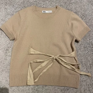 Beige stickad topp från Zara - Säljer en superfin beige stickad topp från Zara. Den har korta ärmar och en snygg knytning i midjan som ger en extra touch. Perfekt för både skola och middag