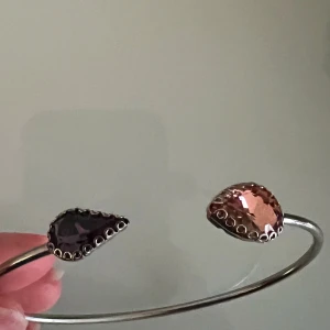 Silverarmband med lila och rosa stenar - Snyggt och stilrent silverarmband med två stora droppformade stenar i lila och rosa. Perfekt för att ge en färgklick till din outfit. Armbandet är öppet och justerbart, vilket gör det enkelt att ta på och av. Passar både till vardags och festliga tillfällen!