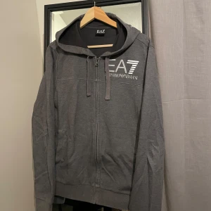Grå hoodie från Emporio Armani - Säljer en snygg grå hoodie från Emporio Armani i storlek S passar även M. Den har en dragkedja framtill och en logga på bröstet. Perfekt för en casual look och funkar även som unisex. Den är i bra skick och superbekväm att ha på sig. 