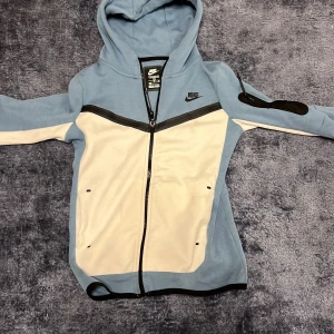 Blå och vit hoodie från Nike - Säljer en snygg blå och vit hoodie från Nike i mycket bra skick. Den har en dragkedja framtill och en huva för extra mysfaktor. Perfekt för höst och vår, med en cool design och bekväm passform. Passar både till träning och vardagsoutfits!
