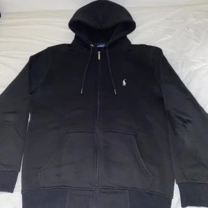 Svart Ralph Lauren Kofta - Säljer en snygg svart kofta från Ralph Lauren i mycket fint skick. Den har dragkedja framtill och en klassisk logga på bröstet. Perfekt för höst och vinter, med en skön passform och praktiska fickor. Huvan är justerbar med snören. Passar både till vardags och chill.