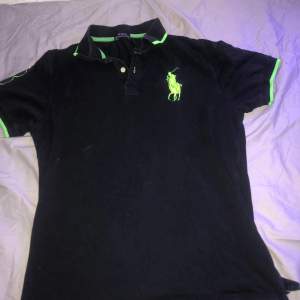 Säljer en snygg svart Polo Ralph Lauren t-shirt i storlek L. Den har coola gröna detaljer och en stor broderad logga på bröstet. Perfekt för en avslappnad stil eller en dag på stan. T-shirten är i bra skick och redo för nya äventyr!