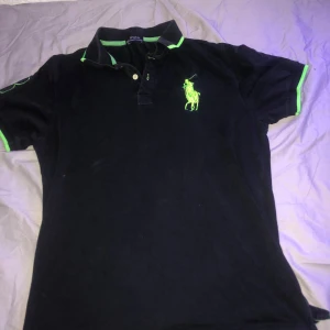 Svart Polo Ralph Lauren t-shirt med gröna detaljer - Säljer en snygg svart Polo Ralph Lauren t-shirt i storlek L. Den har coola gröna detaljer och en stor broderad logga på bröstet. Perfekt för en avslappnad stil eller en dag på stan. T-shirten är i bra skick och redo för nya äventyr!