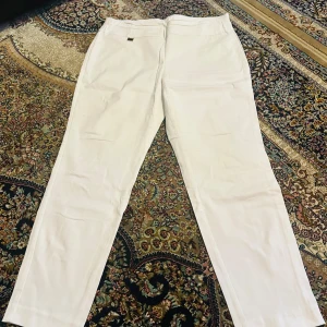 Vita byxor från Alfani - Säljer ett par stilrena vita byxor från Alfani i storlek 14S. De är gjorda av en mjuk och stretchig blandning av rayon, nylon och spandex, vilket gör dem bekväma att bära. Perfekta för både vardag och festliga tillfällen. Byxorna har en klassisk design med raka ben.