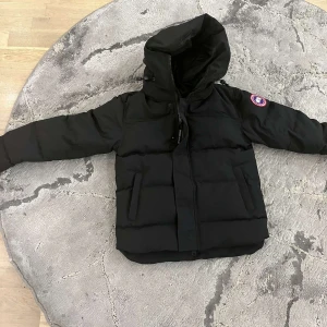 Svart dunjacka från Canada Goose - Säljer en svart dunjacka från Canada Goose i mycket bra skick. Jackan har en stor huva och en dragkedja framtill. Perfekt för kalla vinterdagar. Den har en klassisk design med en logga på ärmen. Supervarm och stilren!