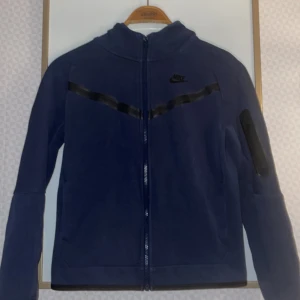 Mörkblå tech fleece från Nike - Säljer en mörkblå tech fleece från JD Sports i fint skick har använt några gånger inga hål är i storlek 147-158. Nypris 1000 mitt pris 500. Tröjan har två fickor vid magen utan dragkedja men har också en liten ficka i ärmen som visas i bild 4. 