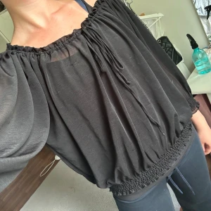 Svart off-shoulder blus - Säljer en svart blus med trekvartsärmar. Blusen har en lös passform och är tillverkad i ett lätt och luftigt material. Den har en knytning framtill vid halsringningen och elastiska band vid ärmslut och nederkant. Perfekt för både vardag och fest!