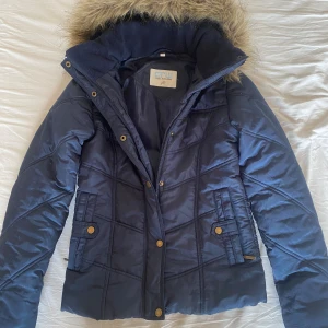 Blå vinterjacka från CRW - Säljer en snygg och varm blå vinter rid jacka från CRW. Jackan har en mysig päls på huvan och är quiltad för extra värme. Den har både dragkedja och knappar framtill för att hålla kylan ute. Perfekt för kalla vinterdagar! ❤️