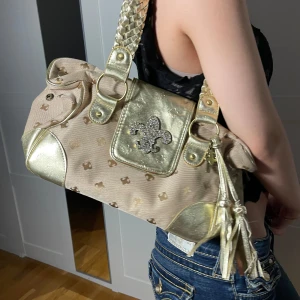 Beige och guld axelväska med tofsar - Säljer en snygg beig och guld väska från Friis and company.Väskan har en flik med en glittrig detalj och tofsar som hänger ner. två rhinestones har gått av men annars av bra skick💕
