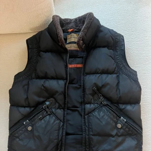 Svart quiltad dunväst från Parajumpers - Säljer en svart quiltad dunväst från Parajumpers i storlek herr M. Västen har en dragkedja framtill och två fickor med dragkedja och knappar. Insidan är fodrad med mjuk fleece för extra värme. Perfekt för höst och vinter!