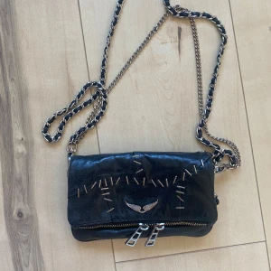 Svart axelväska från Zadig & Voltaire - Säljer en snygg svart axelväska från Zadig & Voltaire. Väskan är gjord av skinn och har en cool design med silverfärgade dragkedjor och detaljer. Den har en kedjerem som kan bäras över axeln eller crossbody. Perfekt för både vardag och fest!