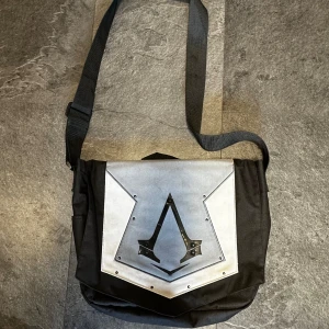 Svart axelväska med Assassin's Creed-logga - Säljer en svart axelväska med en stor Assassin's Creed-logga på framsidan. Väskan har ett justerbart axelband och flera fack inuti, inklusive ett med dragkedja. Perfekt för att bära med sig böcker, laptop eller andra vardagssaker.