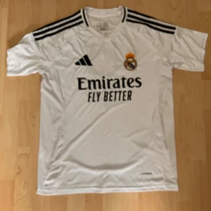Vit Real Madrid fotbollströja med Mbappe #6 - Säljer en vit Real Madrid fotbollströja från Adidas med Mbappe #9 på ryggen. Tröjan har korta ärmar och är i nyskick. Den har svarta detaljer. Perfekt för alla Real Madrid-fans! Lite suddiga bilder men ifall ni vill ha bättre bilder får ni komma privat, tack!