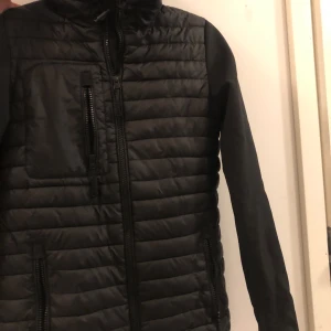 Svart pufferjacka med huva. Skriv för mer bilder, priset kan diskuteras vid snabb affär. ☺️ - Säljer en svart pufferjacka från crossover. Använd ett par gånger, nästa. helt ny. Jackan har en quiltad design med dragkedja framtill och en praktisk huva. Den har långa ärmar och en ficka med dragkedja på bröstet. Perfekt för kyliga dagar!                                           Ny pris är 1723 kr