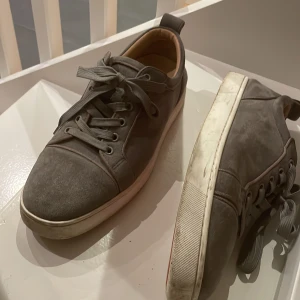 Grå sneakers med röd sula - Snygga grå sneakers med snörning och en unik röd sula (självklart äkta) Skorna har en stilren design med en bekväm passform, perfekt för vardagsbruk. De är tillverkade i mocka och har en vit sula som ger en fräsch kontrast. Lite smutsig sula men kan rengöras hur enkelt som helst så den blir skinande.