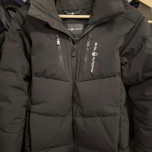 Svart dunjacka från Sail Racing - Sail Racing vinterjacka, varm, vindtät och vattenavvisande. Några små, knappt synliga hål. 