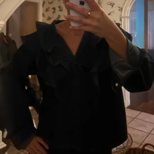 Denimskjorta med volangdetaljer - Snygg svart jacka med volangdetaljer och knappar framtill. Jackan har långa ärmar och en unik design, priset kan diskuteras💕