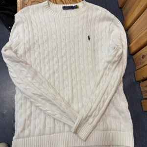 Vit kabelstickad tröja från Ralph Lauren - Säljer en stilren vit kabelstickad tröja från Ralph Lauren. Tröjan har en klassisk rund halsringning och det ikoniska logotypbroderiet på bröstet. Perfekt för en elegant och avslappnad look. (OBS) koftan har en liten fläck som kan gå bort i tvätten 