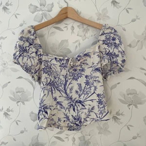 Blommig korsettopp med puffärmar - Charmig korsettopp med blått blommönster på vit bakgrund. Toppen har puffiga kortärmar och en söt knytdetalj framtill. Perfekt för en somrig look!