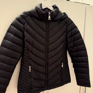 Svart dunjacka från Calvin Klein - Snygg svart dunjacka från Calvin Klein med dragkedja och huva. Jackan har en quiltad design och långa ärmar. Perfekt för kyligare dagar. Som ny