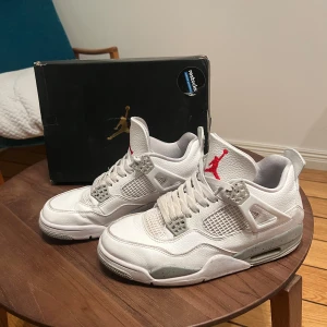 Vita Nike Air Jordans - Snygga vita Nike Air Jordans med grå och röda detaljer. Skorna har en klassisk design med snörning och perforerade paneler för ventilation. Perfekta för den som vill ha en stilren och sportig look.