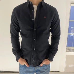 Svart skjorta från Burberry - Skjorta från Burberry i bomull. Knappen längst ner är defekt se bild 5. Bortsett från det är den i mycket bra skick. Storlek medium. Modellen är 180cm och väger 75kg. 