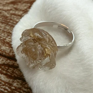 Ring med blomma i genomskinligt material - Unik ring med en stor blomma i genomskinligt material och guldfolie, som ger en elegant och stilren look. Ringen har en silverfärgad bas som kompletterar blomman perfekt. Perfekt för att ge en extra touch till din outfit. Ringen är justerbar