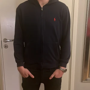  Ralph Lauren marin blå hoodie M/S - Säljer en marin blå hoodie från Ralph Lauren med dragkedja och det klassiska röda logotypen på bröstet. Perfekt för en avslappnad look.