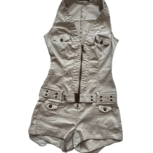 Beige jumpsuit från Jane Norman - Snygg beige jumpsuit från Jane Norman med dragkedja framtill och bälte i midjan. Den har knappar på bröstfickorna och en stilren krage. Perfekt för en avslappnad och trendig look.