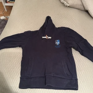Mörkblå hoodie Hackett - Storlek xl men sitter som en M. Säljer en mörkblå hoodie med ett stilrent emblem på bröstet. Hoodien har en klassisk design med snörning i halsen och en stor ficka framtill. Perfekt för en avslappnad stil.
