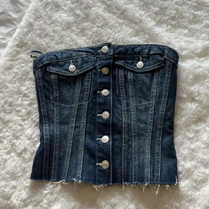 Jeanskorsett med knappar - Snygg jeanskorsett med knappar framtill och dekorativa fickor. Den har en elastisk rygg för bättre passform och råa kanter för en trendig look. Perfekt för att styla med olika outfits.