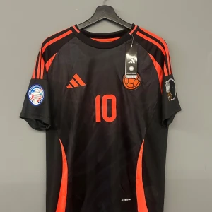 James Rodriguez Colombia 24/25 helt ny - Snygg svart fotbollströja från Adidas med röda detaljer och nummer 10 på både fram- och baksidan. Tröjan har korta ärmar och är dekorerad med Copa America-märken på ärmarna. Perfekt för fotbollsfans som vill sticka ut på planen! Den är storlek M och är en replika