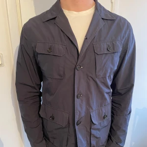Marinblå overshirt/fieldjacket från J.Lindeberg - Snygg mörkblå jacka från J.Lindeberg med knappar framtill och fyra fickor. Perfekt för en stilren look. Jackan är knappt använd och är tillverkad i ett lätt material.
