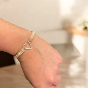Beige armband med hjärtdetalj - Charmigt beige armband med en öppen hjärtdetalj i silverfärg. Armbandet har en flätad design som ger en bohemisk känsla. Perfekt för att lägga till en subtil touch av stil till din outfit.