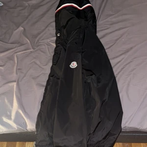 Svart jacka från Moncler - Säljer en stilren svart jacka från Moncler med en diskret logotyp på ärmen. Jackan har en klassisk design med dragkedja och en randig detalj vid kragen. Perfekt för dig som vill ha en snygg och funktionell jacka.