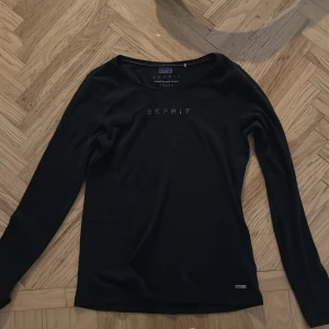 Svart långärmad topp från Esprit - Säljer en stilren svart långärmad topp från Esprit med märkeslogga i små stenar på bröstet. Perfekt för en enkel och elegant look. Passar bra till både jeans och kjol.