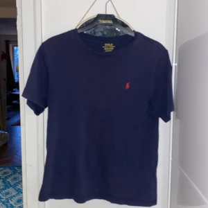 Mörkblå t-shirt från Polo Ralph Lauren - Säljer en mörkblå t-shirt från Polo Ralph Lauren med det klassiska röda logotypbroderiet på bröstet. T-shirten är kortärmad och tillverkad i bomull, perfekt för en avslappnad stil. Tröjan har några få små hål som man ser på tredje bilden och är lite urtvättad.