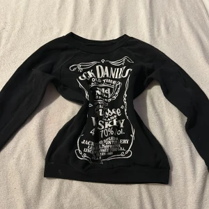Svart tröja med Jack Daniels-tryck - Snygg svart tröja med ett stort vitt Jack Daniels-tryck på framsidan. Perfekt för den som gillar en rockig stil. Tröjan har långa ärmar och en rund halsringning.