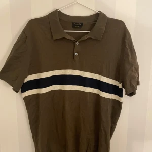 Brun pikétröja från Massimo Dutti - Snygg brun pikétröja från Massimo Dutti med en vit och marinblå rand över bröstet. Tröjan har korta ärmar och en klassisk krage med knappar. Perfekt för en stilren look. Tveka inte att hojta till vid eventuella frågor!