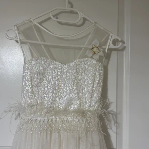 Vit långklänning med paljetter - Elegant vit långklänning med glittrande paljetter upptill och transparenta axelband. Klänningen har en vacker blomdetalj vid axeln och fjäderliknande dekorationer i midjan. Perfekt för speciella tillfällen. Behövs bara tvätt 