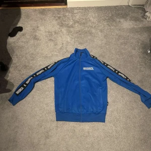 Hoodrich hoodie  - Säljer en blå track jacket från Hoodrich med dragkedja och textdetaljer längs ärmarna. Jackan har en hög krage och ribbade muddar vid ärmslut och nederkant. Perfekt för en avslappnad stil.