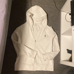 Vit hoodie från Ralph Lauren - Snygg vit hoodie från Ralph Lauren med dragkedja och klassisk logga på bröstet. Perfekt för en avslappnad stil. Den har en huva med snörning och praktiska fickor framtill.