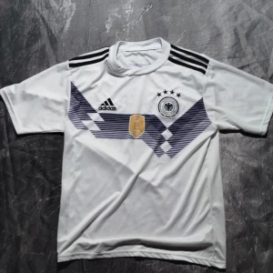 Vit fotbollströja från Adidas - Säljer en vit fotbollströja från Adidas med svarta detaljer och tre stjärnor ovanför ett emblem. Tröjan har korta ärmar och ett tryck med 'Müller' och nummer 13 på ryggen. Perfekt för fotbollsfans!