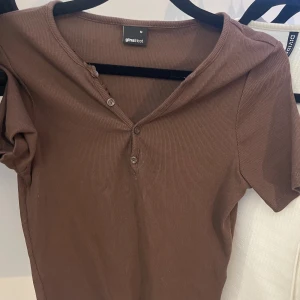 Brun ribbad topp från Gina Tricot - Säljer en brun ribbad topp från Gina Tricot med knappar framtill. Toppen har en rund hals och korta ärmar, perfekt för en avslappnad stil.