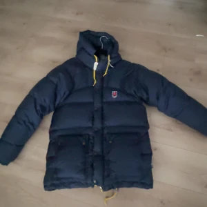 Blå dunjacka från Fjällräven - Jackan är i storlek XXXS men sitter som en S eller M 🤩Jackan är i Navy blue. Blå dunjacka från Fjällräven med huva och gula detaljer. Inga fläckar eller hål. Jackan har knappar och dragkedja framtill. Perfekt för kalla dagar!☺️