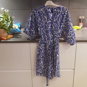 Blå och vit klänning från Lindex - Snygg blå och vit mönstrad klänning från Lindex med knappar framtill och ett knytband i midjan. Klänningen har korta ärmar och ett unikt abstrakt mönster som ger en modern touch.