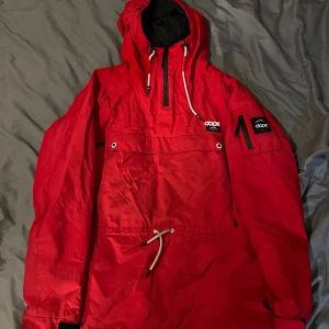Röd vindjacka från Dope - Säljer en röd vindjacka från Dope med justerbar huva och snörning i midjan. Jackan har en stor ficka framtill och märkeslogga på ärmen. Lite små fläckar men annars super fint skick, hela dragkedjor