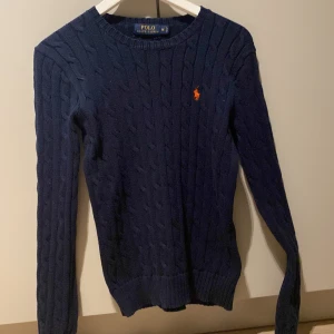 Mörkblå stickad tröja från Polo Ralph Lauren - Snygg mörkblå stickad tröja från Polo Ralph Lauren med det klassiska broderade logotypen i orange på bröstet. Tröjan har en kabelstickad design och långa ärmar, perfekt för kyligare dagar men funkar även på hösten och våren med en väst typ. De är i 9,5/10 i skicket. Den är använd sparsamt. Hör av er om ni har frågor eller prisförslag! 