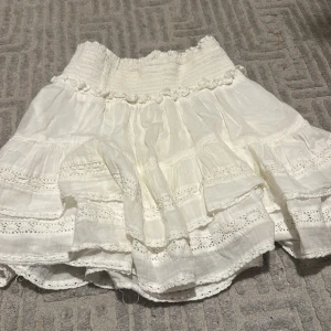 Vit kjol med spetsdetaljer - Charmig vit kjol med volanger och spetsdetaljer. Den har en elastisk midja för bekväm passform och flera lager som ger en luftig känsla. Perfekt för en somrig look!