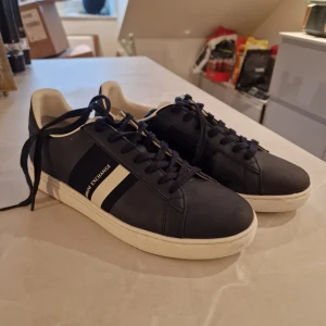 Svarta sneakers från Armani Exchange - Snygga svarta sneakers från Armani Exchange med vita detaljer och snörning. Perfekta för en stilren look. Skorna har en vit sula och A|X-logga på hälen.           ( Endast testade inomhus)